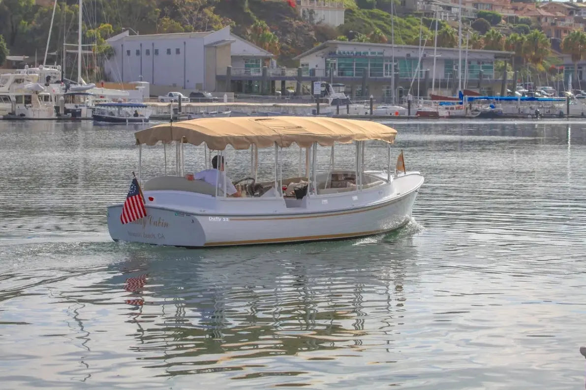 Duffy Cuddy Cabin 22 Electric Boat with a canopy on a calm body of water with buildings and boats in the background.