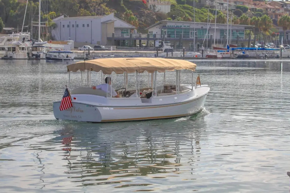 Duffy Cuddy Cabin 22 Electric Boat with a canopy on a calm body of water with buildings and boats in the background.