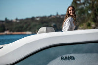Woman standing on Rand Spirit 25 Electric Boat with 'RAND' branding, water and trees in the background